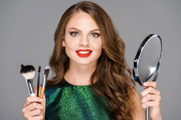 Portrait de belle femme souriante avec pinceaux de maquillage et miroir isolé sur gris — Photo de stock