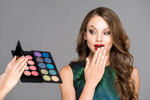 Partial view of makeup artist showing eyeshadows palette to shocked model isolated on grey — Stock Photo