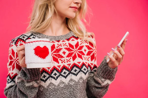 Обрізане зображення дівчини в светрі, що тримає чашку з серцевим знаком і використовує смартфон ізольовано на рожевому — стокове фото