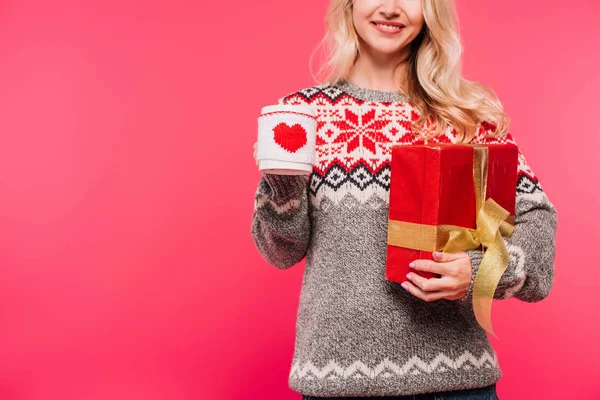 Cropped image of smiling woman in sweater holding cup and gift box isolated on pink — Stock Photo