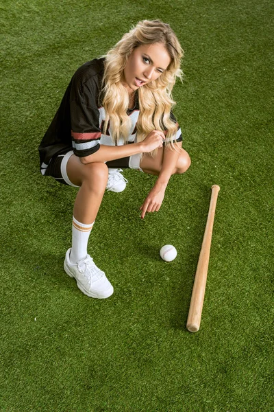 Vista de ángulo alto de la joven atlética en uniforme de fútbol americano con bate de béisbol sentado en la hierba y mirando a la cámara - foto de stock