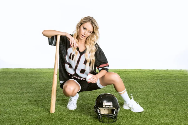 Atlética joven en uniforme de fútbol americano con casco y bate de béisbol sentado en la hierba aislado en blanco - foto de stock