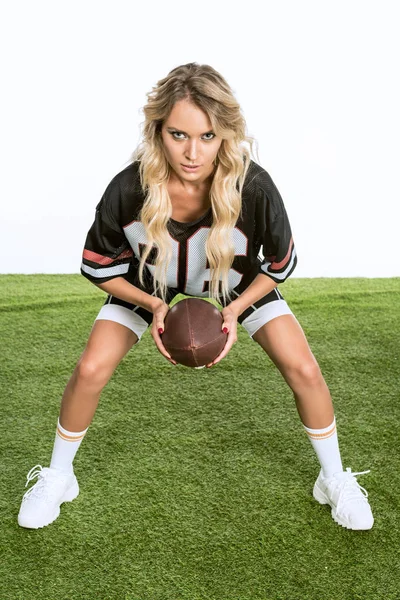 Mujer joven y seria en uniforme de fútbol americano sosteniendo la pelota mientras está de pie sobre hierba verde aislado en blanco - foto de stock
