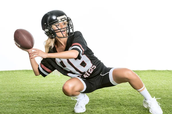 Athletische junge Frau in American-Football-Uniform wirft Ball, während sie hockt hockt auf Gras isoliert auf weiß — Stockfoto