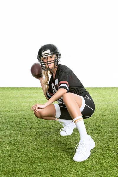 Deportista joven mujer en americano fútbol uniforme lanzando pelota mientras sentado sentadillas en hierba aislado en blanco - foto de stock