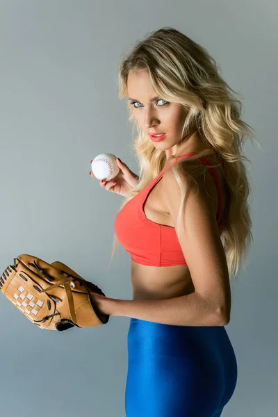Крупним планом портрет привабливої молодої жінки в спортивному одязі з бейсбольною рукавичкою і м'ячем дивиться на камеру на сірому — стокове фото