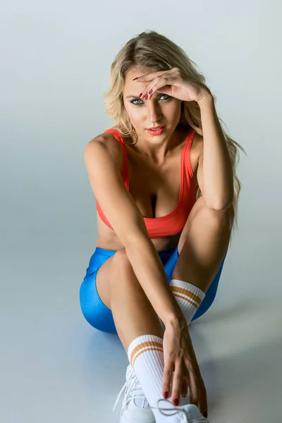 Приваблива спортивна молода жінка дивиться на камеру, сидячи на підлозі на сірому — стокове фото