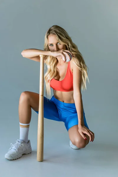 Спортивная молодая женщина с бейсбольной битой и мячом, стоящая на коленях на сером — стоковое фото