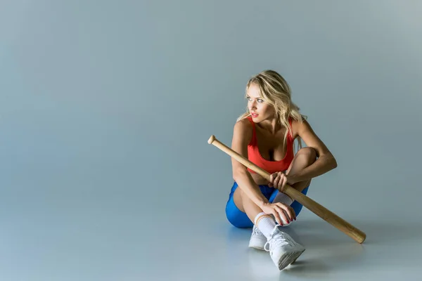 Apto jovem mulher com bastão de beisebol sentado no cinza e olhando para longe — Fotografia de Stock