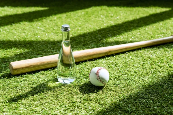 Glass water battle with baseball ball and bat lying on green grass — Stock Photo