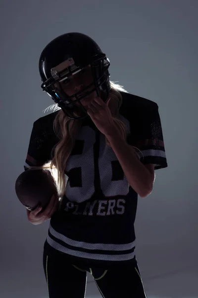 Dark silhouette of sporty young woman in american football equipment with ball on grey — Stock Photo