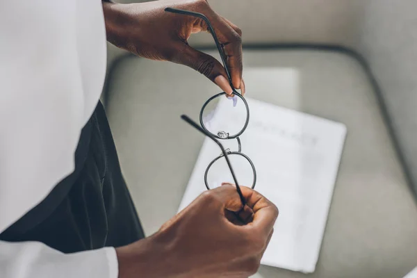 Imagem recortada de empresária afro-americana segurando óculos perto de poltrona no escritório — Fotografia de Stock
