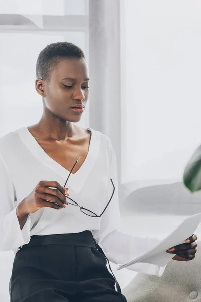 Elegante mujer de negocios afroamericana leyendo documentos y sosteniendo gafas en la oficina - foto de stock