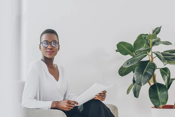 Стильна приваблива афроамериканська бізнес-леді сидить у кріслі з документами в офісі — стокове фото