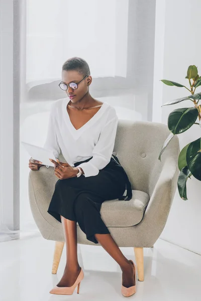 Mujer de negocios afroamericana atractiva de moda sentada en sillón y leyendo documentos en la oficina - foto de stock