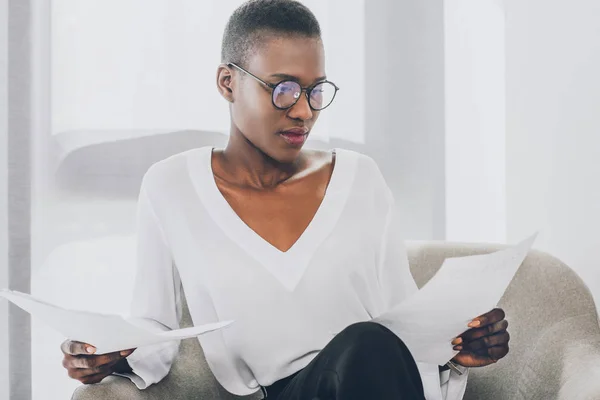 Elegante atractiva mujer de negocios afroamericana sentada en sillón y leyendo documentos en la oficina - foto de stock