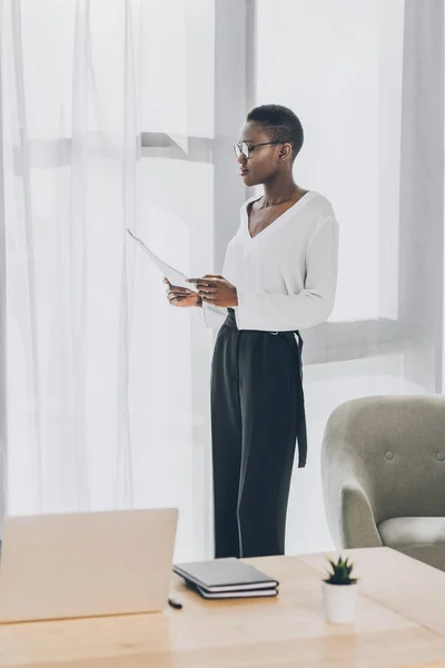 Elegante atractiva mujer de negocios afroamericana de pie y leyendo documentos en la oficina — Stock Photo