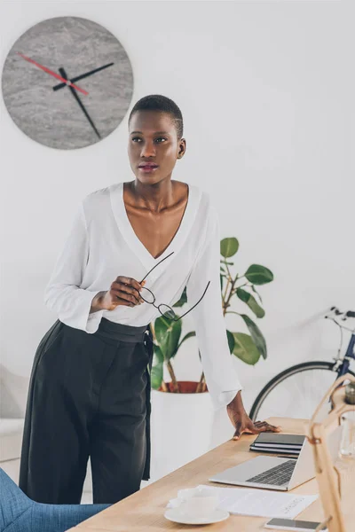 Модна приваблива афроамериканська бізнес-леді тримає окуляри і спирається на стіл в офісі — стокове фото