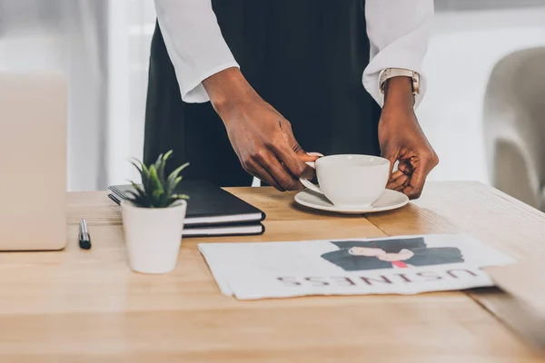 Обрізане зображення стильної афроамериканської бізнес-леді, що кладе чашку кави на стіл в офісі — стокове фото