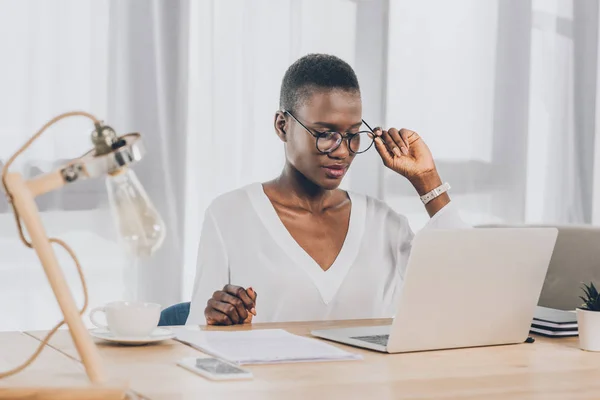Стильна приваблива афроамериканська бізнес-леді торкається окулярів і дивиться на ноутбук в офісі — стокове фото
