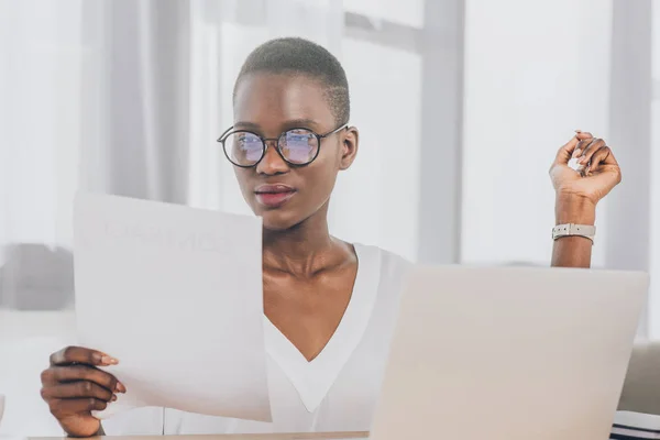 Стильна приваблива афроамериканська бізнес-леді читає документи за столом в офісі — стокове фото