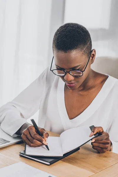 Stylish attractive african american businesswoman writing something to notebook in office — Stock Photo