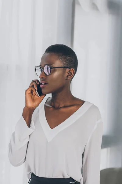 Elegante atractiva mujer de negocios afroamericana hablando por teléfono inteligente en la oficina y mirando hacia otro lado - foto de stock