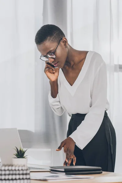 Elegante atractiva mujer de negocios afroamericana hablando por teléfono inteligente y mirando el ordenador portátil en la oficina - foto de stock