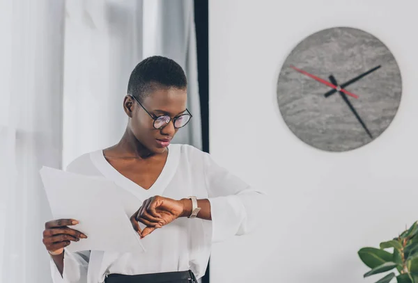 Elegante mujer de negocios afroamericana sosteniendo documentos y comprobando el tiempo en la oficina, reloj en la pared blanca - foto de stock