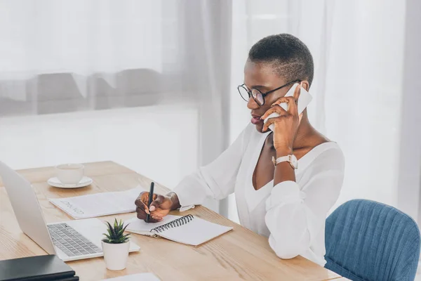 Stylische attraktive afrikanisch-amerikanische Geschäftsfrau spricht per Smartphone und schreibt Notizen im Büro — Stockfoto
