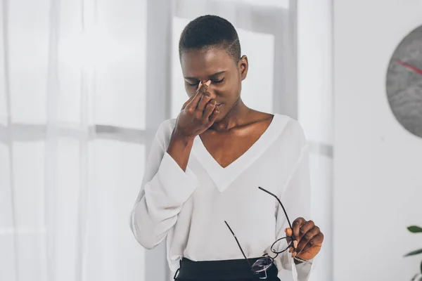 Tired stylish african american businesswoman touching nose bridge and holding glasses in office — Stock Photo