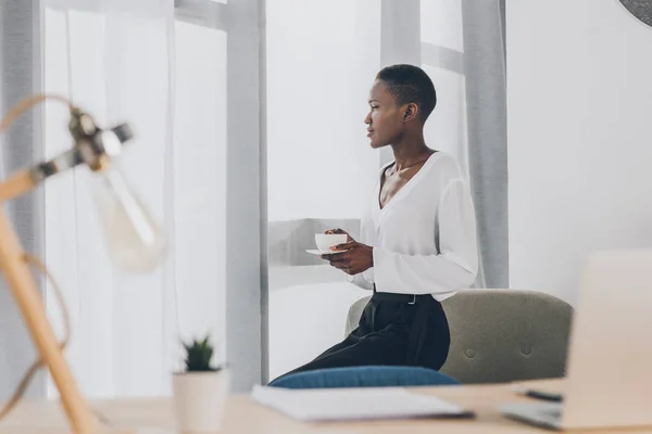 Side view of stylish african american businesswoman sitting on armchair and holding cup of coffee in office — Stock Photo