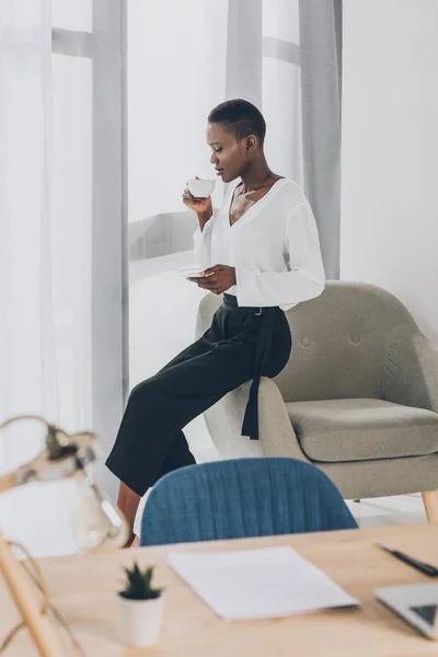 Side view of stylish african american businesswoman sitting on armchair and drinking coffee in office — Stock Photo