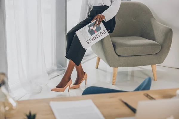 Cropped image of african american businesswoman sitting on armchair and holding newspaper in office — Stock Photo