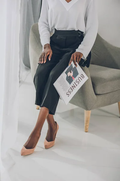 Imagem cortada de mulher de negócios americana africana elegante sentado em poltrona e segurando jornal no escritório — Fotografia de Stock