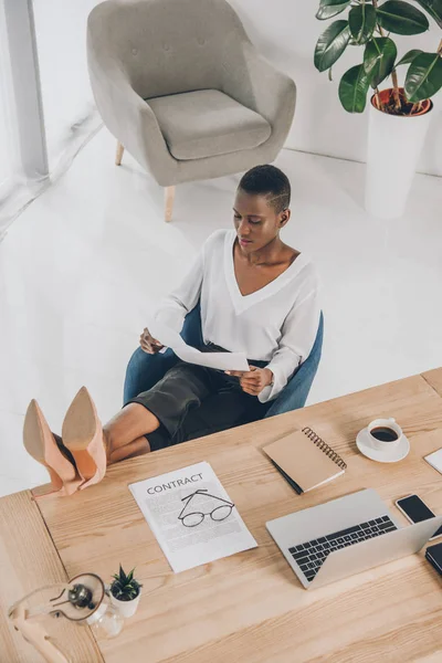 Vista de ángulo alto de elegante atractiva mujer de negocios afroamericana lectura de documentos con patas en la mesa en la oficina - foto de stock