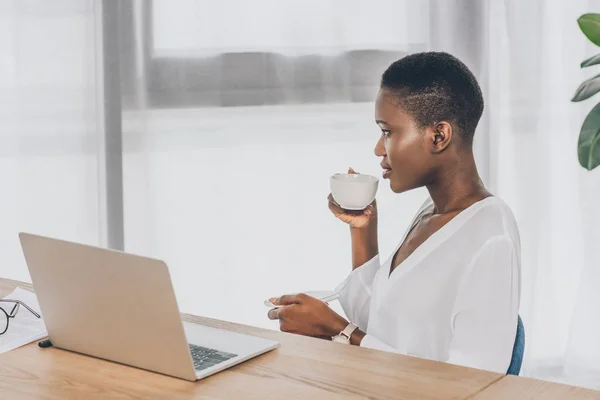 Боковой вид стильной привлекательной африканской бизнесвумен, пьющей кофе в офисе — стоковое фото