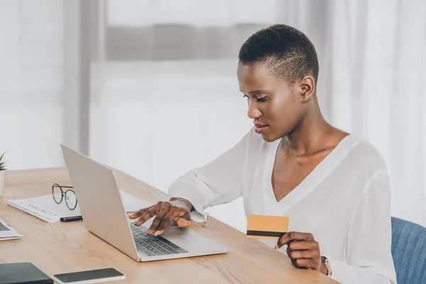 Hermosa mujer de negocios afroamericana de compras en línea con computadora portátil y tarjeta de crédito en la oficina - foto de stock