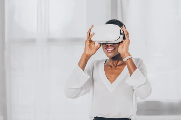 Sonriente mujer de negocios afroamericana elegante en auriculares de realidad virtual en la oficina - foto de stock