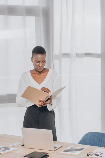 Elegante atractiva mujer de negocios afroamericana buscando en la carpeta en la oficina - foto de stock