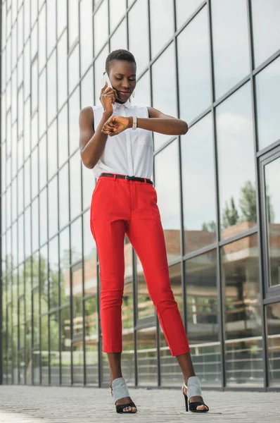 Stylische attraktive afrikanisch-amerikanische Geschäftsfrau, die per Smartphone spricht und die Uhrzeit in der Nähe des Business Centers überprüft — Stockfoto