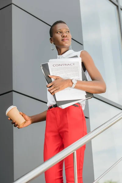 Vue à angle bas d'une femme d'affaires afro-américaine élégante tenant un café à emporter et regardant loin près du bâtiment — Photo de stock