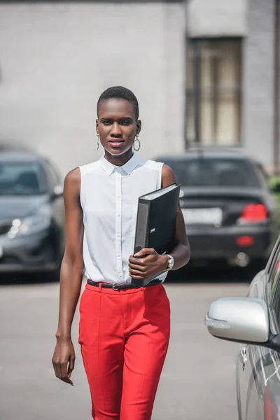 Elegante atractiva mujer de negocios afroamericana caminando cerca de coche en el aparcamiento - foto de stock