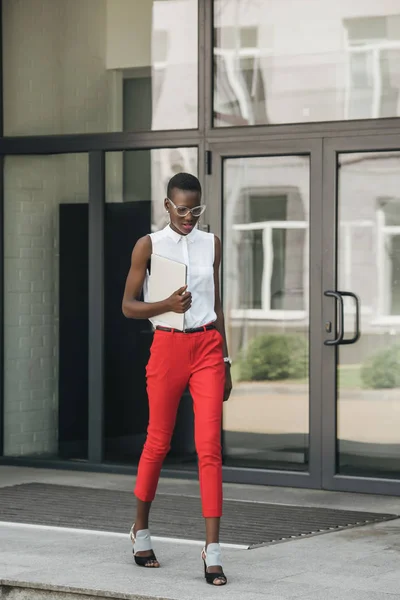 Mujer de negocios afroamericana atractiva de moda caminando con el ordenador portátil en la calle - foto de stock