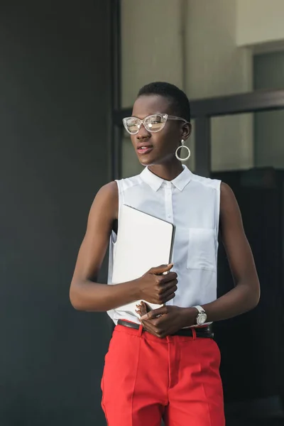 Élégant attrayant afro-américain femme d'affaires tenant ordinateur portable et regardant loin dans la rue — Photo de stock