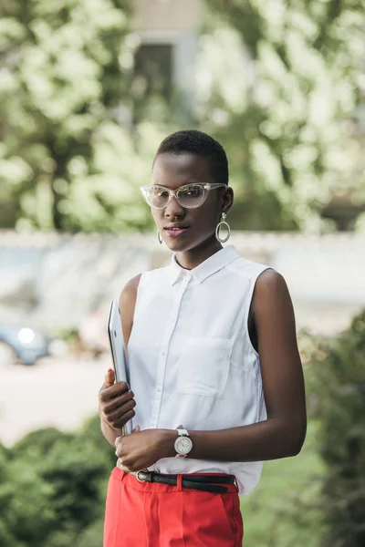 Приваблива афроамериканська бізнес-леді в модному одязі, дивлячись на камеру і тримаючи ноутбук на вулиці — стокове фото