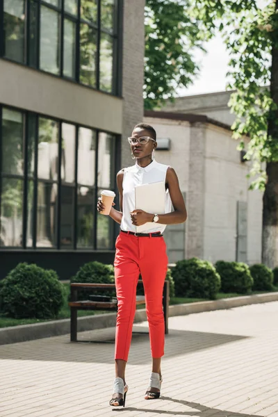 Élégante femme d'affaires afro-américaine attrayante marchant dans la rue avec café dans une tasse en papier et ordinateur portable — Photo de stock