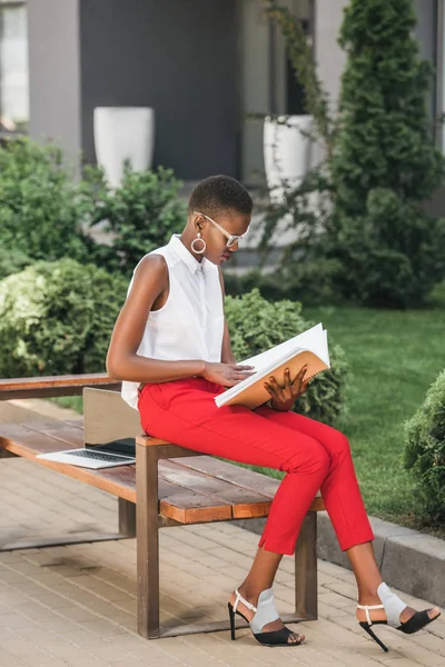 Fashionable african american businesswoman sitting on bench and reading documents on street — Stock Photo