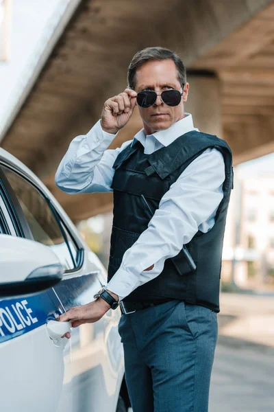 Oficial de policía masculino serio en chaleco antibalas quitándose las gafas de sol cerca del coche en la calle - foto de stock