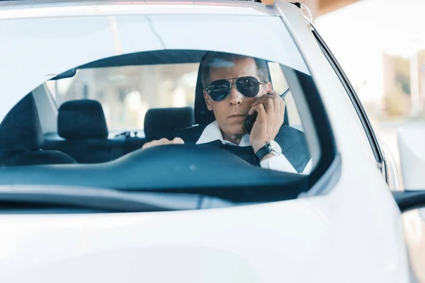 Serious mature policeman in sunglasses talking on radio set in car — Stock Photo
